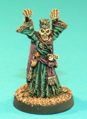 Pose 1, variant A. This is the Lich, an evil spellcaster in Undead form. He wears once-opulent robes and a cloak which are now badly tattered, and has a sceptre, a purse and a couple of rolled scrolls tucked into his sash. This particular variant has a completely skeletal head, and wears a jewelled crown. His mouth is closed and he looks directly forwards.